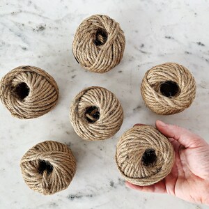 May include: Six balls of natural twine string, each wound into a ball shape. The balls are arranged in a semi-circle on a white marble surface.