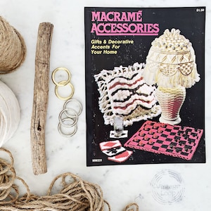 Może przedstawiać: Książka zatytułowana "Macrame Accessories: Gifts & Decorative Accents For Your Home" z ceną 1,50 $. Okładka książki przedstawia różne projekty makramy, w tym abażur, poduszkę i szachownicę. Książka jest otoczona różnymi materiałami do makramy, w tym sznurkiem, włóczką, drewnianym prętem i metalowymi pierścieniami.