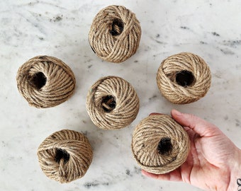 May include: Six balls of natural twine string, each wound into a ball shape. The balls are arranged in a semi-circle on a white marble surface.
