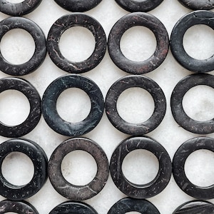 Puede incluir: Una colección de cuentas redondas de madera negra y gris con un acabado liso y pulido. Las cuentas tienen una superficie ligeramente texturizada y están dispuestas en un patrón de cuadrícula.