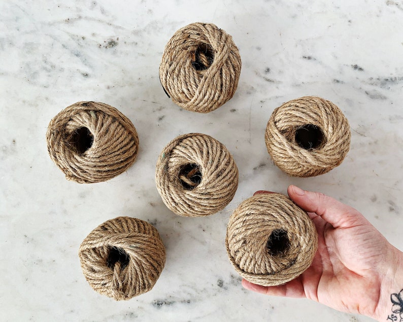 May include: Six balls of natural twine string, each wound into a ball shape. The balls are arranged in a semi-circle on a white marble surface.