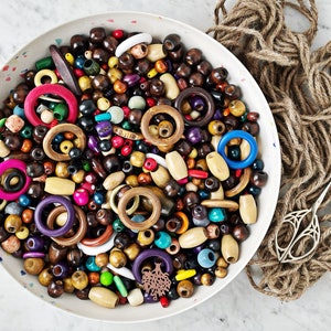 Peut inclure: Un bol blanc rempli de perles en bois de différentes couleurs, notamment des perles rondes, des perles en forme de beignets et des perles longues et cylindriques. Les perles sont disposées de manière aléatoire et présentent un mélange de tons de bois naturels, de couleurs vives et de nuances brun foncé.