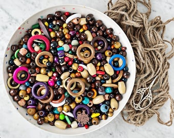 May include: A white bowl filled with a variety of colorful wooden beads, including round beads, donut-shaped beads, and long cylinder beads. The beads are arranged in a random pattern and are a mix of natural wood tones, bright colors, and dark brown shades.