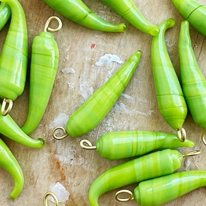 May include: A selection of light green glass chilli pepper charms with gold-coloured loops for jewellery making. The peppers have a striped design and are arranged on a light brown wooden surface.