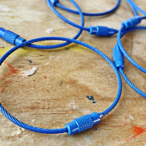 May include: Close-up of blue cable keyrings with metal ends. The keyrings are coiled and lying on a wooden surface.