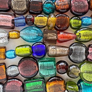 May include: A collection of multi-colored glass beads featuring various shapes including squares, circles, and ovals. The beads display a metallic, foil-like finish in shades of blue, green, orange, red, purple, and silver, with a reflective surface.