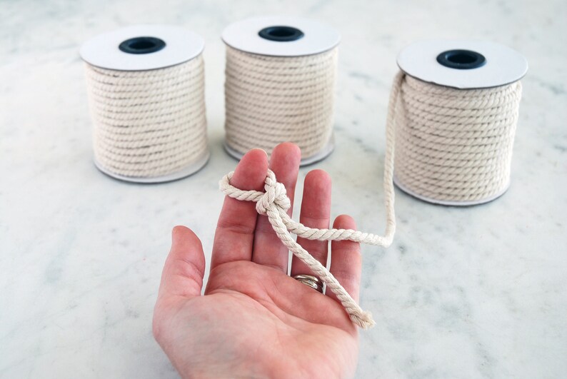 May include: Three spools of natural cotton rope, with one spool being held in a hand, showing a knot tied in the rope.