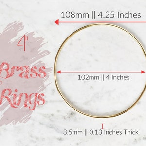 Puede incluir: Un anillo de metal dorado de 102 mm de diámetro y 3,5 mm de grosor. El anillo se muestra sobre una superficie de mármol blanco. El texto "Brass Rings" está escrito en rojo sobre un fondo rosa.