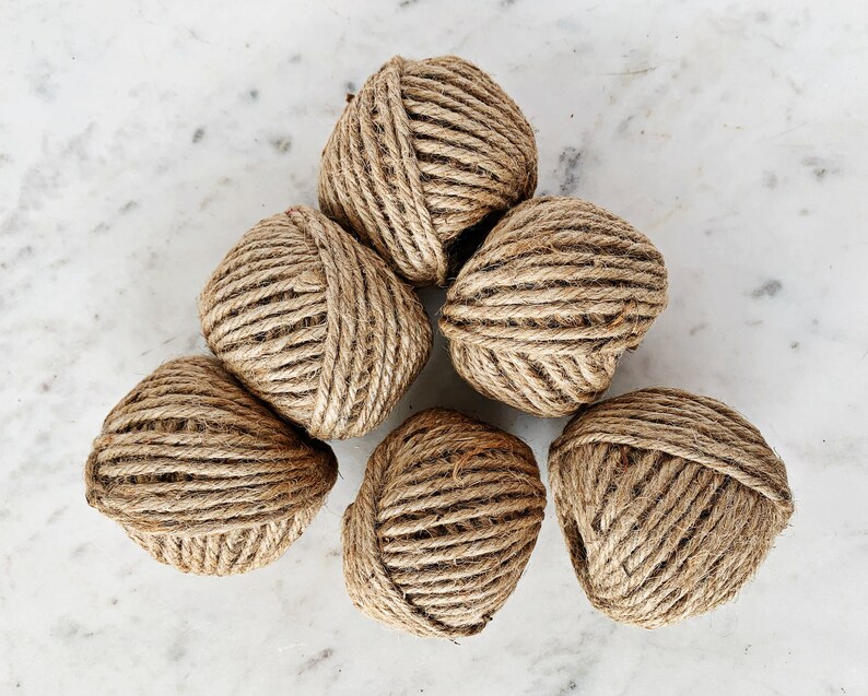 May include: Six balls of natural brown twine, arranged in a triangular shape on a white marble surface.
