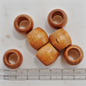 May include: A collection of seven wooden beads with a natural, light brown finish. The beads are cylindrical with a hole running through the center. A ruler is in the image, showing measurements in inches.