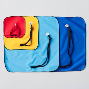 Puede incluir: Cuatro paños de microfibra de diferentes tamaños, rojo, amarillo, azul y azul oscuro, con correas negras y etiquetas blancas. Los paños están doblados y dispuestos en una pila.