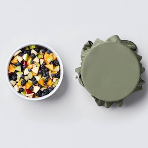 Puede incluir: Un bol blanco lleno de una ensalada de frutas con manzanas, naranjas, arándanos y moras picadas. Un cubre-bol de tela verde con ribete negro se encuentra al lado del bol.