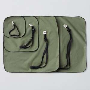 Puede incluir: Cuatro bolsas de tela verde con ribetes negros y correas negras. Las bolsas están anidadas una dentro de la otra, con la bolsa más grande en la parte inferior y la más pequeña en la parte superior.