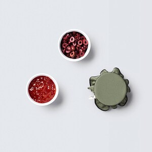 Puede incluir: Tres cuencos blancos sobre un fondo gris. Un cuenco contiene mermelada roja, otro contiene fruta seca roja y marrón, y el tercer cuenco está vacío. Una tapa de tela verde con ribete negro también está en el fondo.