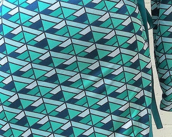 May include: A long-sleeved wrap dress with a geometric pattern in teal, blue, and white. The dress has a V-neck and a tie at the waist. The dress is displayed on a mannequin against a white brick wall.