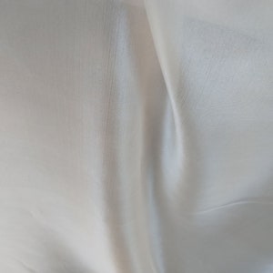 Puede incluir: Primer plano de una tela blanca con un brillo sutil. El material está drapeado, creando pliegues y sombras suaves. La textura parece lisa con un ligero patrón acanalado, lo que sugiere un textil ligero.