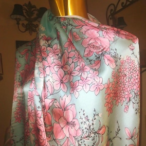 Pode incluir: Um lenço azul-claro com um padrão repetitivo de flores cor-de-rosa e caules castanhos escuros. O tecido tem um acabamento liso e refletor e está drapeado sobre um manequim dourado, exibindo a estampa floral em toda a sua superfície.