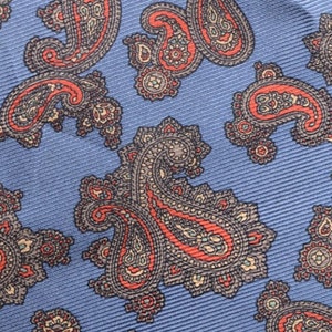 May include: Close-up of a blue fabric with a repeating paisley pattern in shades of red, brown, and beige. The fabric has a subtle ribbed texture, creating a visual depth to the design. Ideal for apparel.