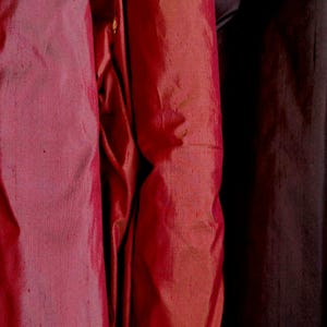 Może przedstawiać: Zbliżenie na trzy drapowane tkaniny w odcieniach czerwieni i bordowego. Tkaniny mają połysk, z widocznymi fałdami i fakturami. Kolory wahają się od jasnoczerwonego do głębokiego, ciemnego bordowego.