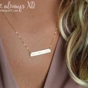 Peut inclure: Un collier dor&eacute; avec une cha&icirc;ne d&eacute;licate. Le pendentif rectangulaire porte des chiffres romains. Le texte "Love always XO" et l'adresse du site web sont dans le coin sup&eacute;rieur gauche.