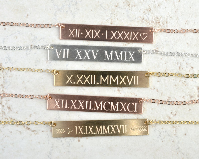 Peut inclure: Plusieurs colliers avec des barres personnalis&eacute;es en or rose, argent et or. Chaque collier est grav&eacute; de chiffres romains et de symboles, avec de fines cha&icirc;nes.