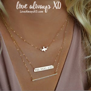 Peut inclure: Trois colliers dor&eacute;s superpos&eacute;s. Le collier sup&eacute;rieur a un pendentif en forme d'oiseau. Le collier du milieu a une barre rectangulaire avec des chiffres romains. Le collier inf&eacute;rieur a une barre avec de petites pierres claires. Le texte "love always XO" est visible.