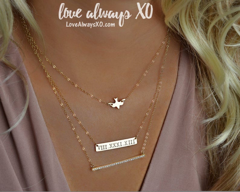 Peut inclure: Trois colliers dor&eacute;s superpos&eacute;s. Le collier sup&eacute;rieur a un pendentif en forme d'oiseau. Le collier du milieu a une barre rectangulaire avec des chiffres romains. Le collier inf&eacute;rieur a une barre avec de petites pierres claires. Le texte "love always XO" est visible.