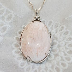 May include: A silver pendant necklace featuring a large, oval-shaped, light pink stone with a feathery pattern. The stone is encased in a silver wire frame and hangs from a silver chain. The background is a white, eyelet-detailed top.