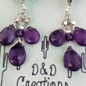 K&ouml;nnte beinhalten: Ein Paar Ohrh&auml;nger mit facettierten, tiefvioletten Amethyst-Tropfen- und runden Perlen. Die Ohrringe sind an silberfarbenen Klappb&uuml;gelverschl&uuml;ssen befestigt. Im Hintergrund ist der Text "D & D Creations" zu sehen.