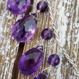 K&ouml;nnte beinhalten: Nahaufnahme einer Halskette mit facettierten, tropfenf&ouml;rmigen Amethyst-Edelsteinen und kleineren runden Amethyst-Perlen. Die violetten Steine sind auf einer silberfarbenen Kette aufgereiht, vor einem rustikalen Holzgrund.