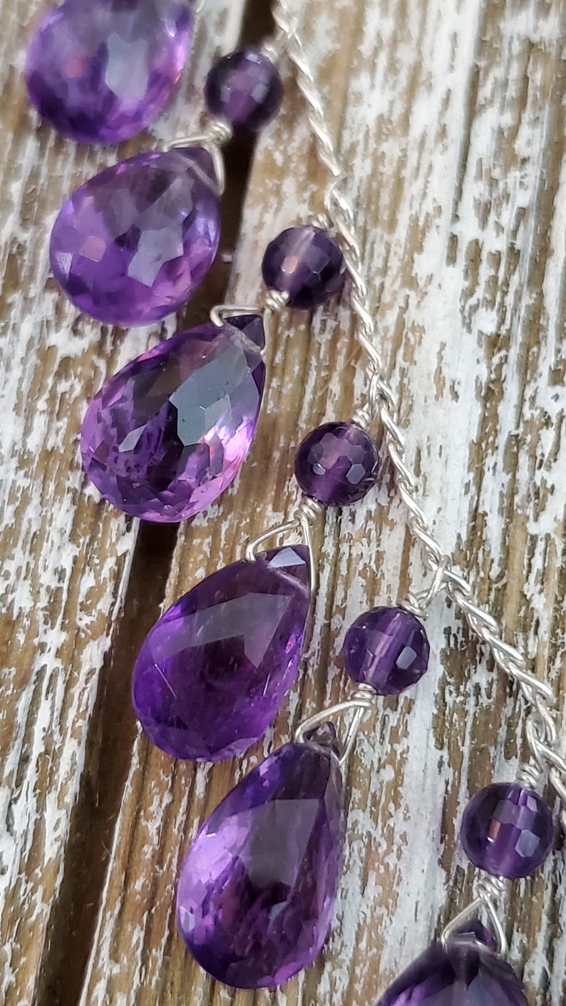 K&ouml;nnte beinhalten: Nahaufnahme einer Halskette mit facettierten, tropfenf&ouml;rmigen Amethyst-Edelsteinen und kleineren runden Amethyst-Perlen. Die violetten Steine sind auf einer silberfarbenen Kette aufgereiht, vor einem rustikalen Holzgrund.