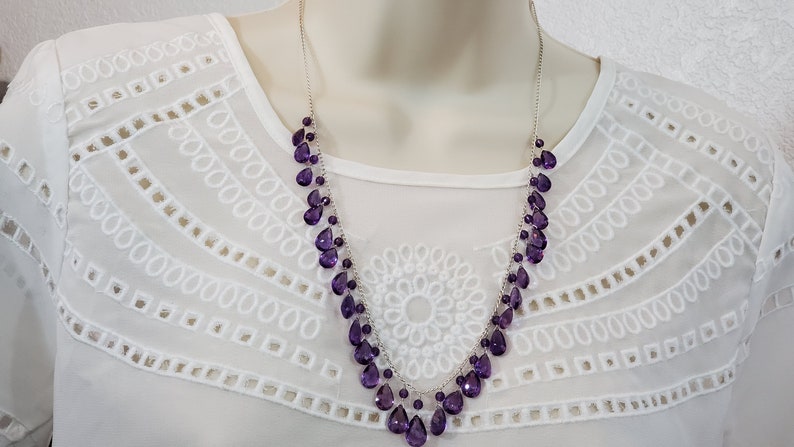 K&ouml;nnte beinhalten: Eine silberne Halskette mit mehreren Str&auml;ngen tropfenf&ouml;rmiger, tiefvioletter Edelsteine. Die Halskette wird vor einer wei&szlig;en, mit &Ouml;sen bestickten Bluse pr&auml;sentiert. Die Edelsteine sind facettiert.