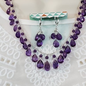 K&ouml;nnte beinhalten: Ein silbernes Halsketten- und Ohrring-Set mit tropfenf&ouml;rmigen, lila Amethyst-Edelsteinen. Die Halskette und die Ohrringe werden auf einer wei&szlig;en Schaufensterpuppe pr&auml;sentiert, um das Design und die Farbe des Schmucks hervorzuheben.