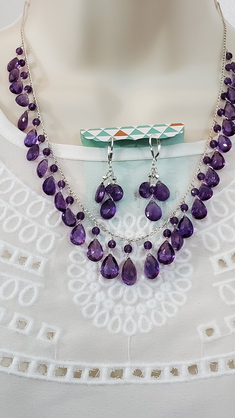K&ouml;nnte beinhalten: Ein silbernes Halsketten- und Ohrring-Set mit tropfenf&ouml;rmigen, lila Amethyst-Edelsteinen. Die Halskette und die Ohrringe werden auf einer wei&szlig;en Schaufensterpuppe pr&auml;sentiert, um das Design und die Farbe des Schmucks hervorzuheben.