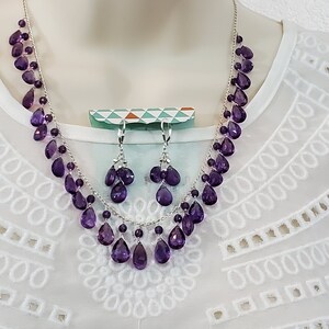 K&ouml;nnte beinhalten: Ein Halsketten- und Ohrring-Set mit tropfenf&ouml;rmigen, tiefvioletten Edelsteinen. Die Halskette und die Ohrringe sind an einer silbernen Kette aufgereiht. Der Schmuck wird vor wei&szlig;em Hintergrund pr&auml;sentiert.