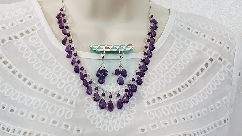 K&ouml;nnte beinhalten: Ein Halsketten- und Ohrring-Set mit tropfenf&ouml;rmigen, tiefvioletten Edelsteinen. Die Halskette und die Ohrringe sind an einer silbernen Kette aufgereiht. Der Schmuck wird vor wei&szlig;em Hintergrund pr&auml;sentiert.