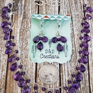 K&ouml;nnte beinhalten: Ein silbernes Halsketten- und Ohrring-Set mit facettierten, lila Amethyst-Edelsteinen. Die Halskette und die Ohrringe werden auf einer Holzoberfl&auml;che mit einer M&uuml;nze als Gr&ouml;&szlig;enreferenz pr&auml;sentiert. Auf einer Karte steht "D & D Creations".