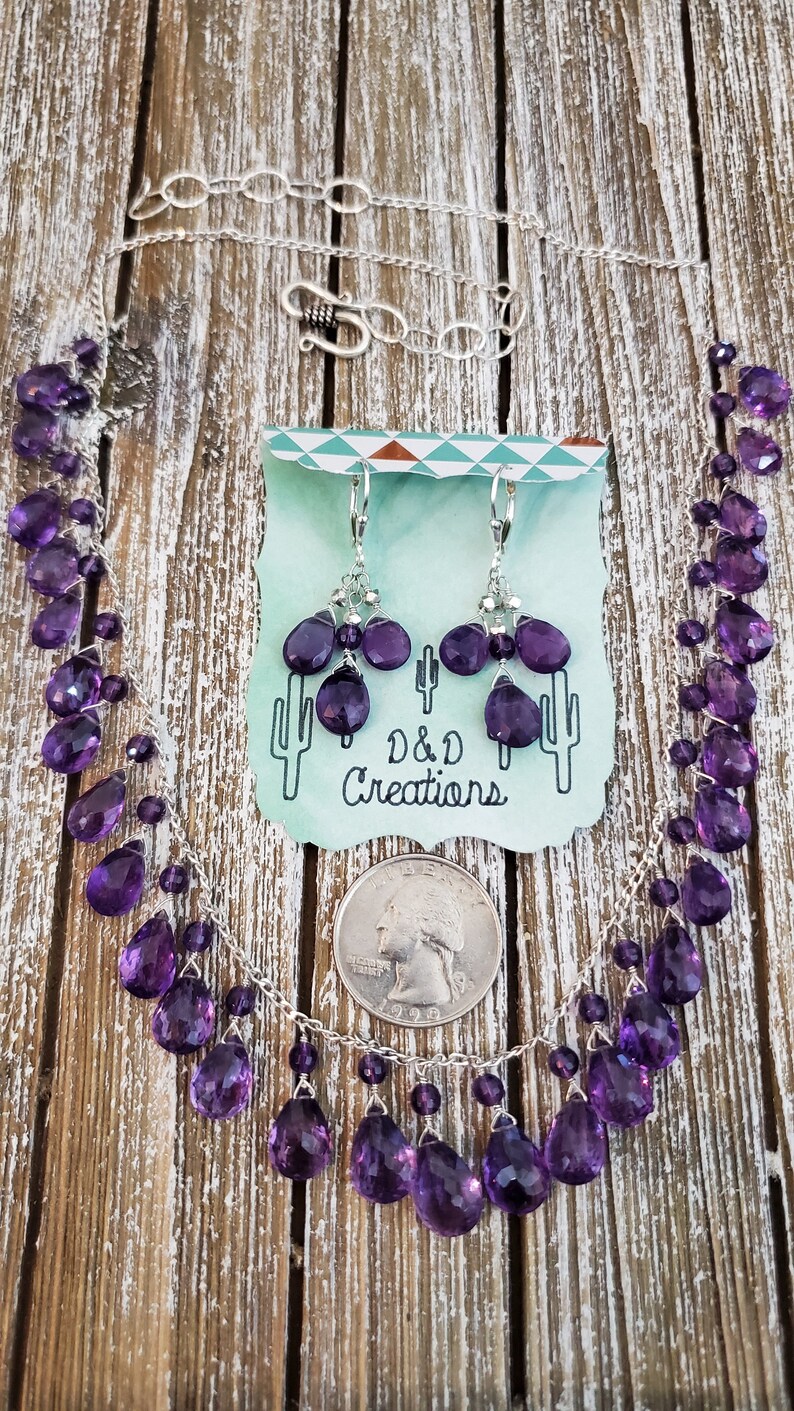 K&ouml;nnte beinhalten: Ein silbernes Halsketten- und Ohrring-Set mit facettierten, lila Amethyst-Edelsteinen. Die Halskette und die Ohrringe werden auf einer Holzoberfl&auml;che mit einer M&uuml;nze als Gr&ouml;&szlig;enreferenz pr&auml;sentiert. Auf einer Karte steht "D & D Creations".