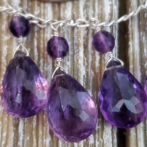 K&ouml;nnte beinhalten: Nahaufnahme einer Silberkette mit Amethyst-Edelsteinanh&auml;ngern. Die Anh&auml;nger sind tropfenf&ouml;rmig und facettiert und weisen eine tiefviolette Farbe auf. Kleine, runde Amethystperlen sind ebenfalls vorhanden. Der Hintergrund ist eine Holzoberfl&auml;che.