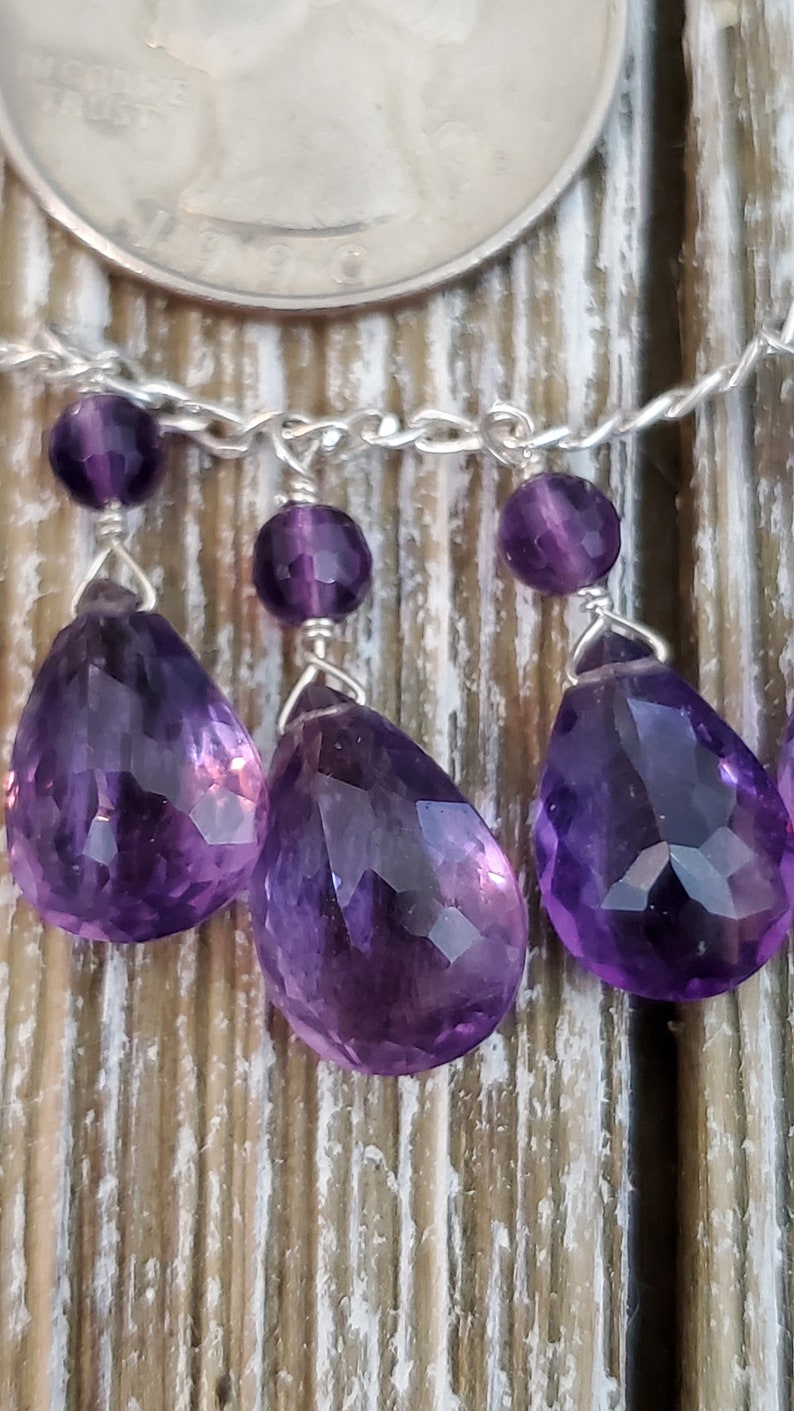K&ouml;nnte beinhalten: Nahaufnahme einer Silberkette mit Amethyst-Edelsteinanh&auml;ngern. Die Anh&auml;nger sind tropfenf&ouml;rmig und facettiert und weisen eine tiefviolette Farbe auf. Kleine, runde Amethystperlen sind ebenfalls vorhanden. Der Hintergrund ist eine Holzoberfl&auml;che.