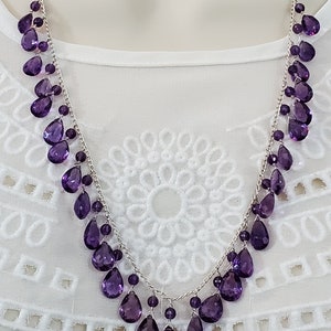 K&ouml;nnte beinhalten: Eine silberne Halskette mit einer Kaskade aus tropfenf&ouml;rmigen, tiefvioletten Edelsteinen. Die Halskette wird vor einem wei&szlig;en Hintergrund pr&auml;sentiert, der das komplizierte Design und die Farbe des Schmucks hervorhebt. Die Edelsteine sind in einem zarten, flie&szlig;enden Muster angeordnet.