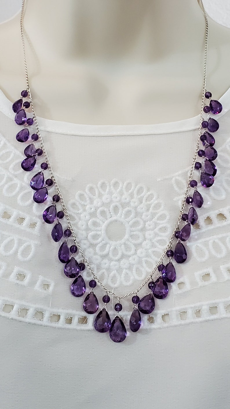 K&ouml;nnte beinhalten: Eine silberne Halskette mit einer Kaskade aus tropfenf&ouml;rmigen, tiefvioletten Edelsteinen. Die Halskette wird vor einem wei&szlig;en Hintergrund pr&auml;sentiert, der das komplizierte Design und die Farbe des Schmucks hervorhebt. Die Edelsteine sind in einem zarten, flie&szlig;enden Muster angeordnet.