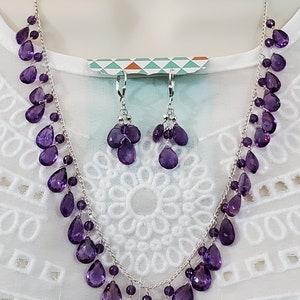 K&ouml;nnte beinhalten: Ein silbernes Halsketten- und Ohrring-Set mit tropfenf&ouml;rmigen, tiefvioletten Edelsteinen. Das Schmuckset wird auf einer wei&szlig;en Oberfl&auml;che pr&auml;sentiert, um das Design und die Farbe hervorzuheben.