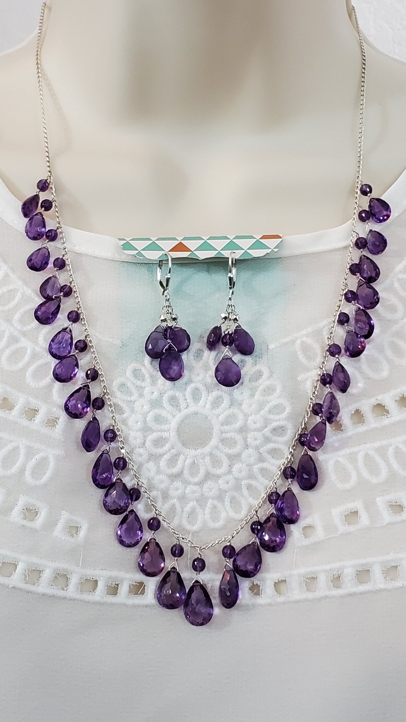 K&ouml;nnte beinhalten: Ein silbernes Halsketten- und Ohrring-Set mit tropfenf&ouml;rmigen, tiefvioletten Edelsteinen. Das Schmuckset wird auf einer wei&szlig;en Oberfl&auml;che pr&auml;sentiert, um das Design und die Farbe hervorzuheben.