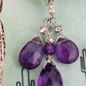 K&ouml;nnte beinhalten: Silberne Ohrh&auml;nger mit facettierten, tiefvioletten Amethyst-Tropfen- und runden Perlen. Die Ohrringe haben einen B&uuml;gelverschluss und sind an einer Silberkette aufgeh&auml;ngt. Der Hintergrund ist hellgr&uuml;n.
