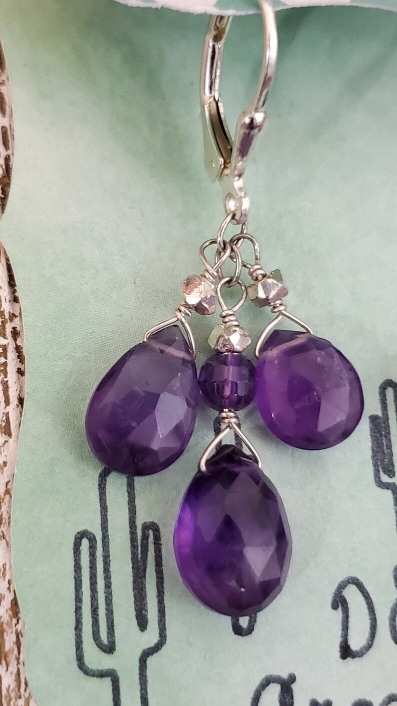 K&ouml;nnte beinhalten: Silberne Ohrh&auml;nger mit facettierten, tiefvioletten Amethyst-Tropfen- und runden Perlen. Die Ohrringe haben einen B&uuml;gelverschluss und sind an einer Silberkette aufgeh&auml;ngt. Der Hintergrund ist hellgr&uuml;n.