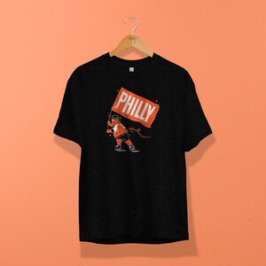 Op de afbeelding: Zwart t-shirt met een afbeelding van een persoon die een oranje vlag vasthoudt met de tekst "PHILLY". Het shirt hangt aan een houten hanger tegen een oranje achtergrond.