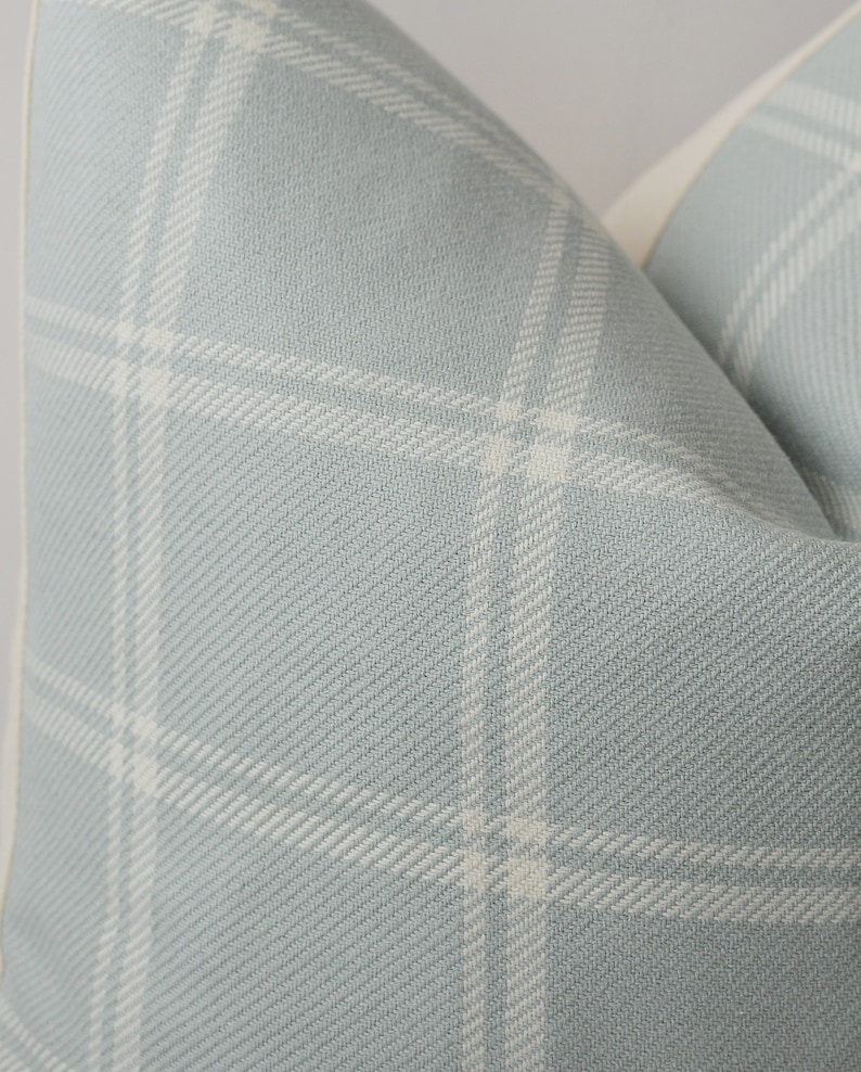 May include: Close-up of a light blue pillow with a white plaid pattern. The pillow has a white border.