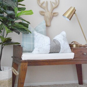 May include: A wooden bench with a white cushion and two pillows, one blue and white plaid and the other white with a gray pattern. A gold lamp and a wooden deer head are on the wall behind the bench.