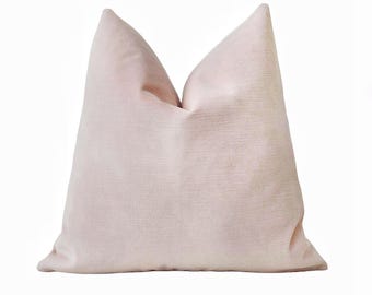 Puede incluir: Un cojín decorativo rosa claro con una superficie texturizada. El cojín, de forma cuadrada, tiene un aspecto suave y afelpado. El cojín es de color liso y está sobre un fondo blanco.